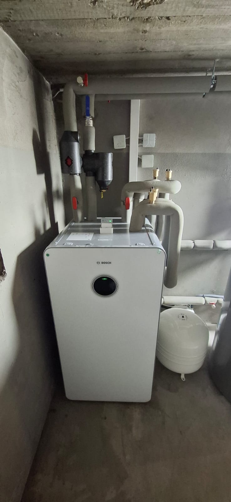 Wärmepumpen-Inneneinheit mit Pufferspeicher in grauem Technikraum, isolierte Rohre verlaufen an Wand und Decke.
